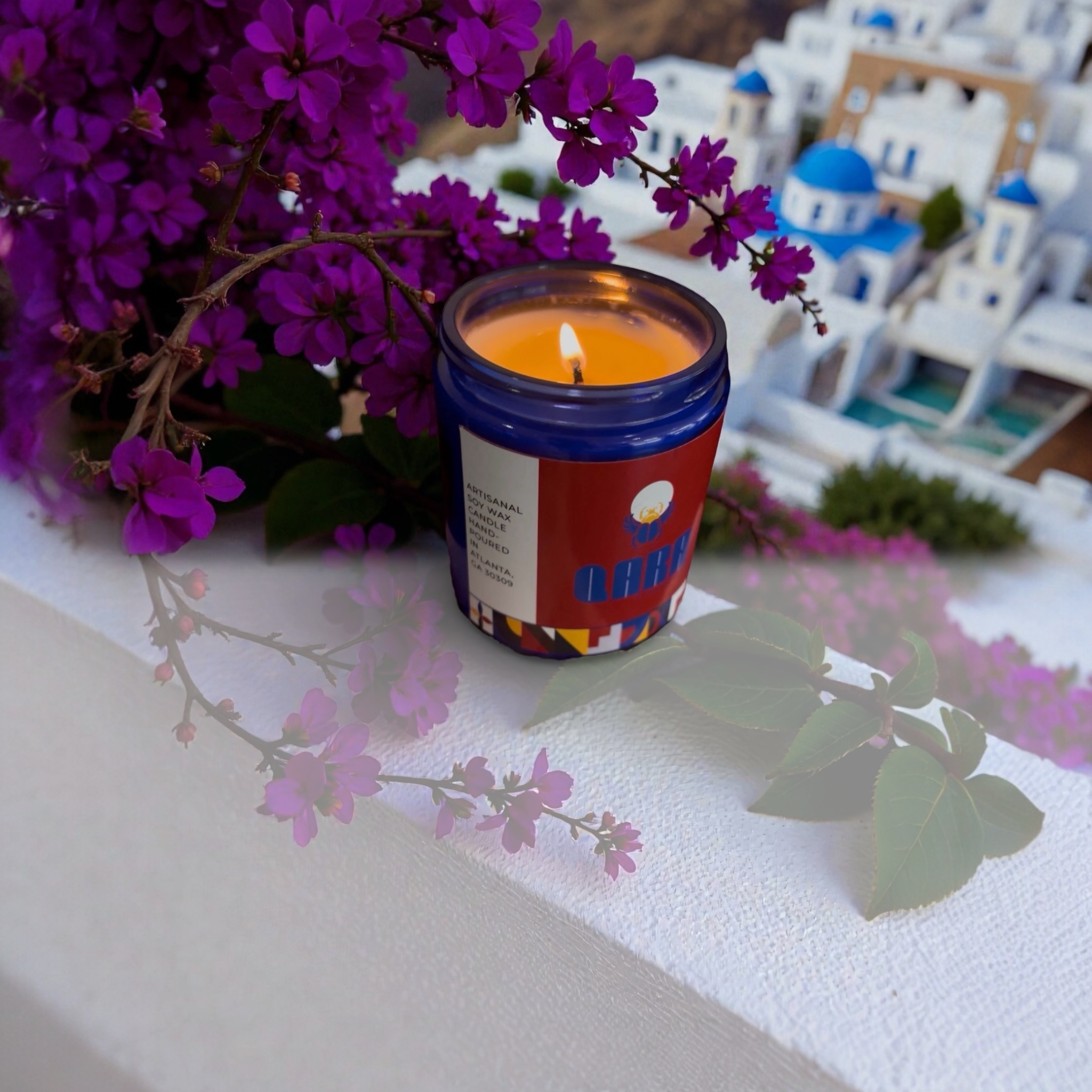 Candle in a colorful container with purple flowers and a blurred background of buildings.