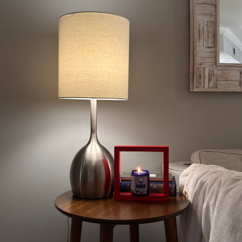 Lamp on a bedside table with a candle and decorative items in a bedroom Candle jar with a purple label featuring the brand 'Qara' on a beige background. QARA IVORY ORIS scented candle Violet Fig  flowery scent luxurious scent natural ingredients and lab tested fragrance oil front cobalt blue jar