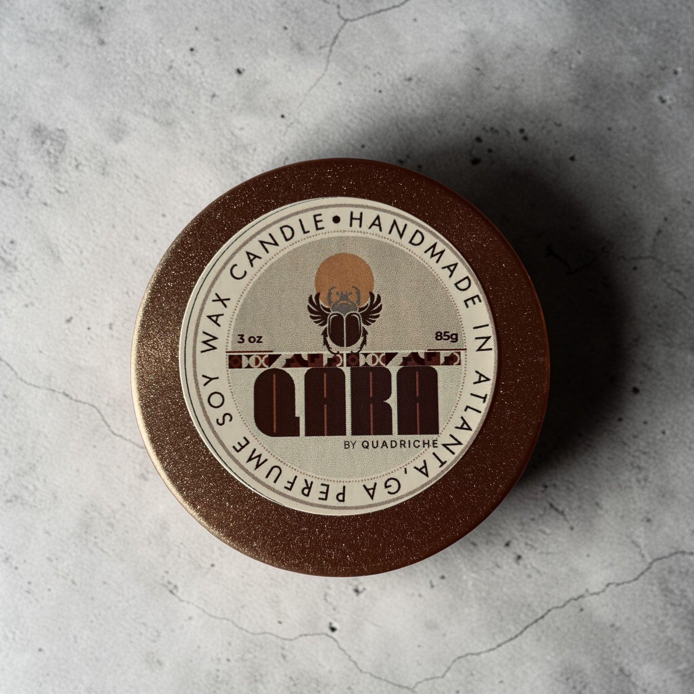 Soy wax candle in a discovery travel round gold tin container with 'QARA by Quadriche' branding on a textured gray surface