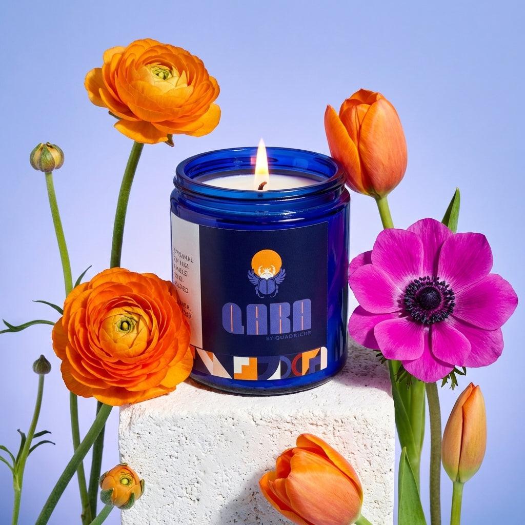 Candle in a blue jar with a label, surrounded by orange and pink flowers on a light blue background
