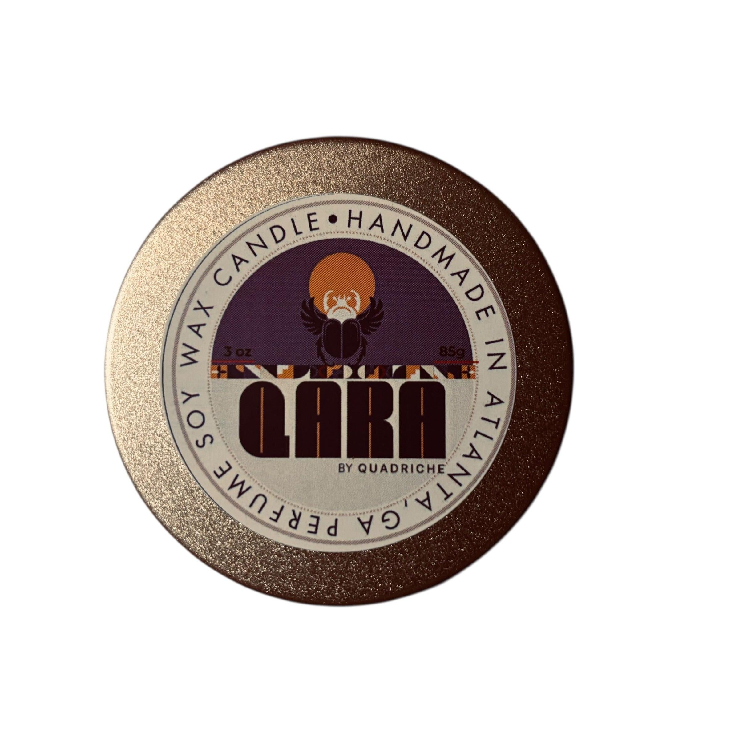 Ember Ink QARA by Quadriche Discovery Travel 3oz Candle in a champagne gold tin with a label on a concrete surface peppercorn violet pine sage juniper leather fragrance scent