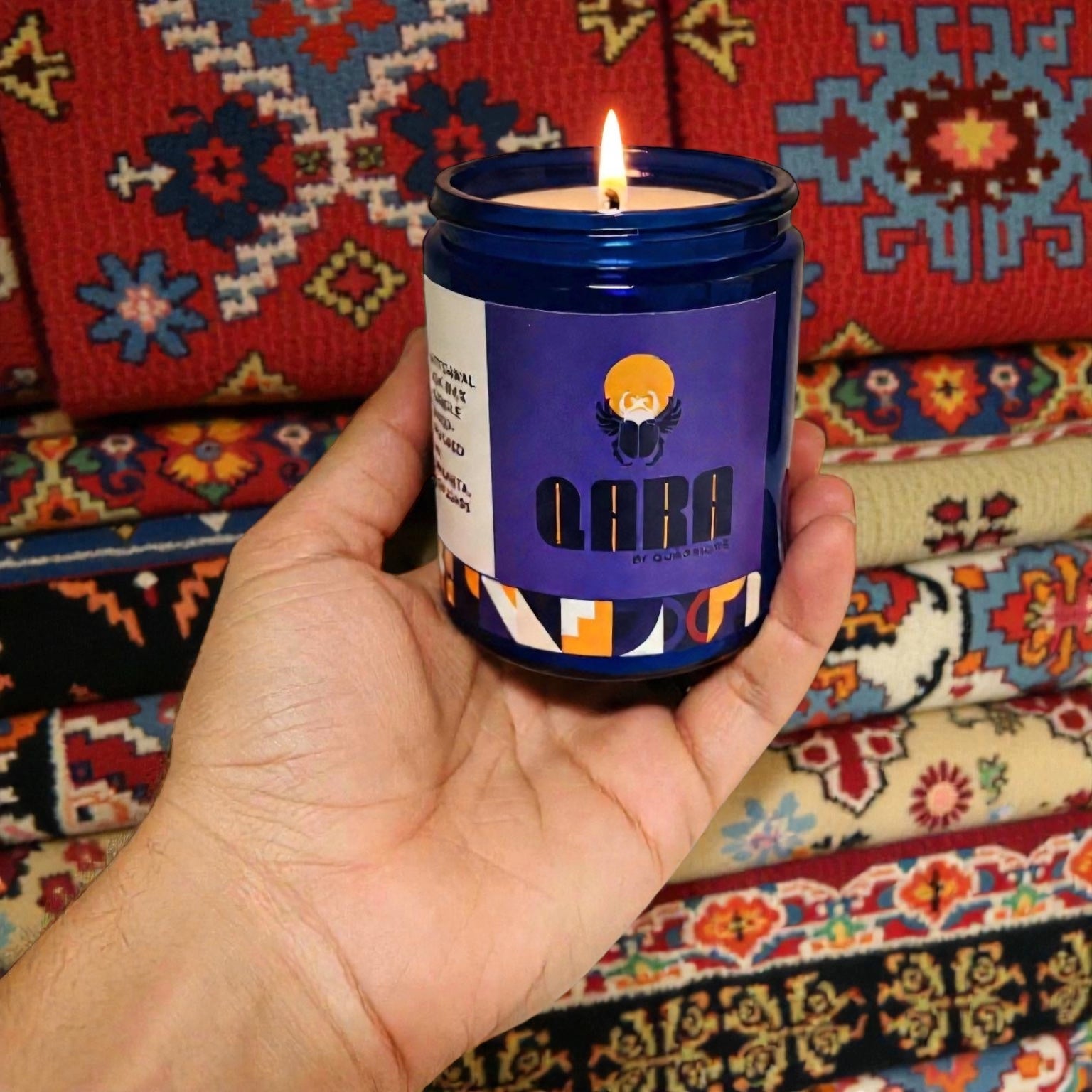 Hand holding a lit candle in a blue container with a colorful label against a Morrocan patterned fabric background