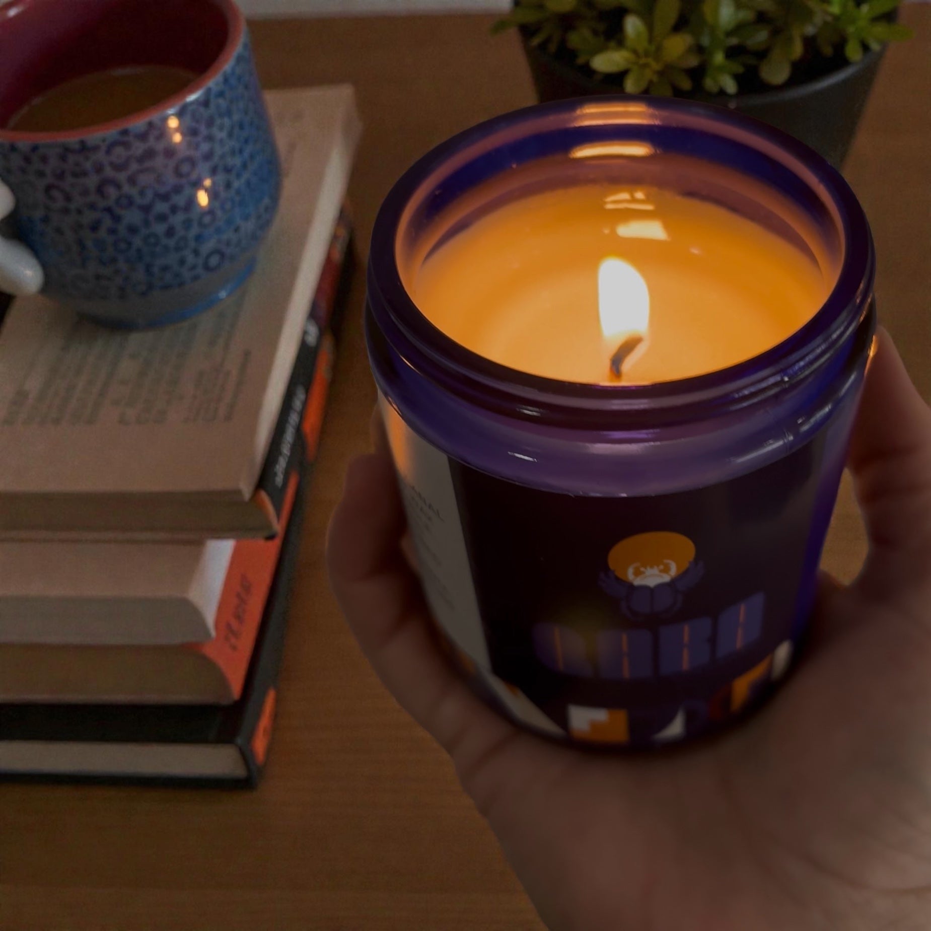Hand holding a lit candle in a purple container with a blurred background of books and a mug.