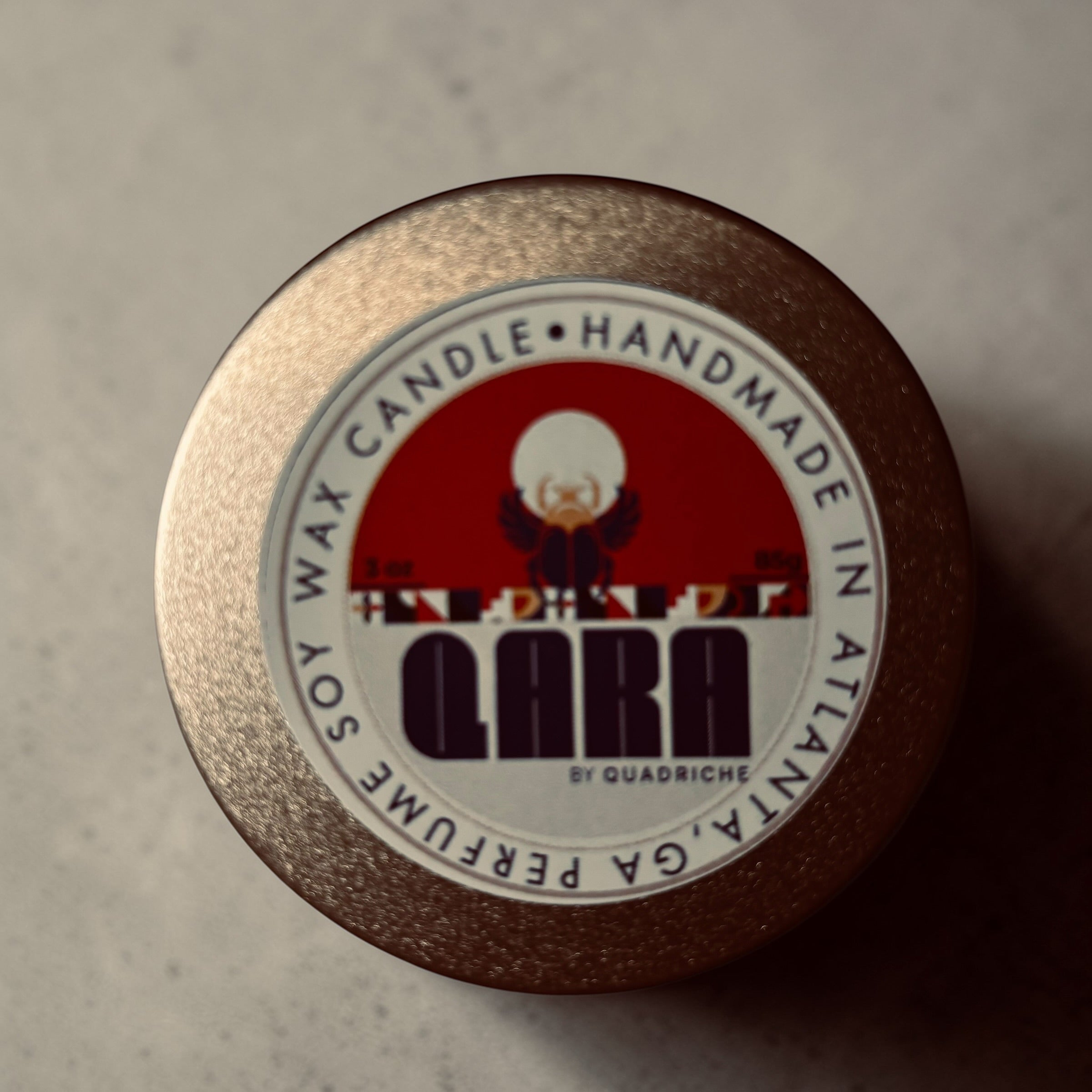 Close-up of aQuadriche coconut soy wax candle label on a textured surface in a champagne gold tin 3oz discovery travel candle