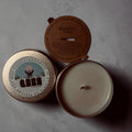 Scented candle with a lid showing branding on a gray surface Dew Discovery Travel 3oz Candle in a tin with a label on a wooden surface ozone apple eucalyptus scent 