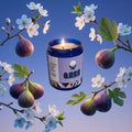 Candle in a blue jar with a label, surrounded by figs and flowers on a clear background