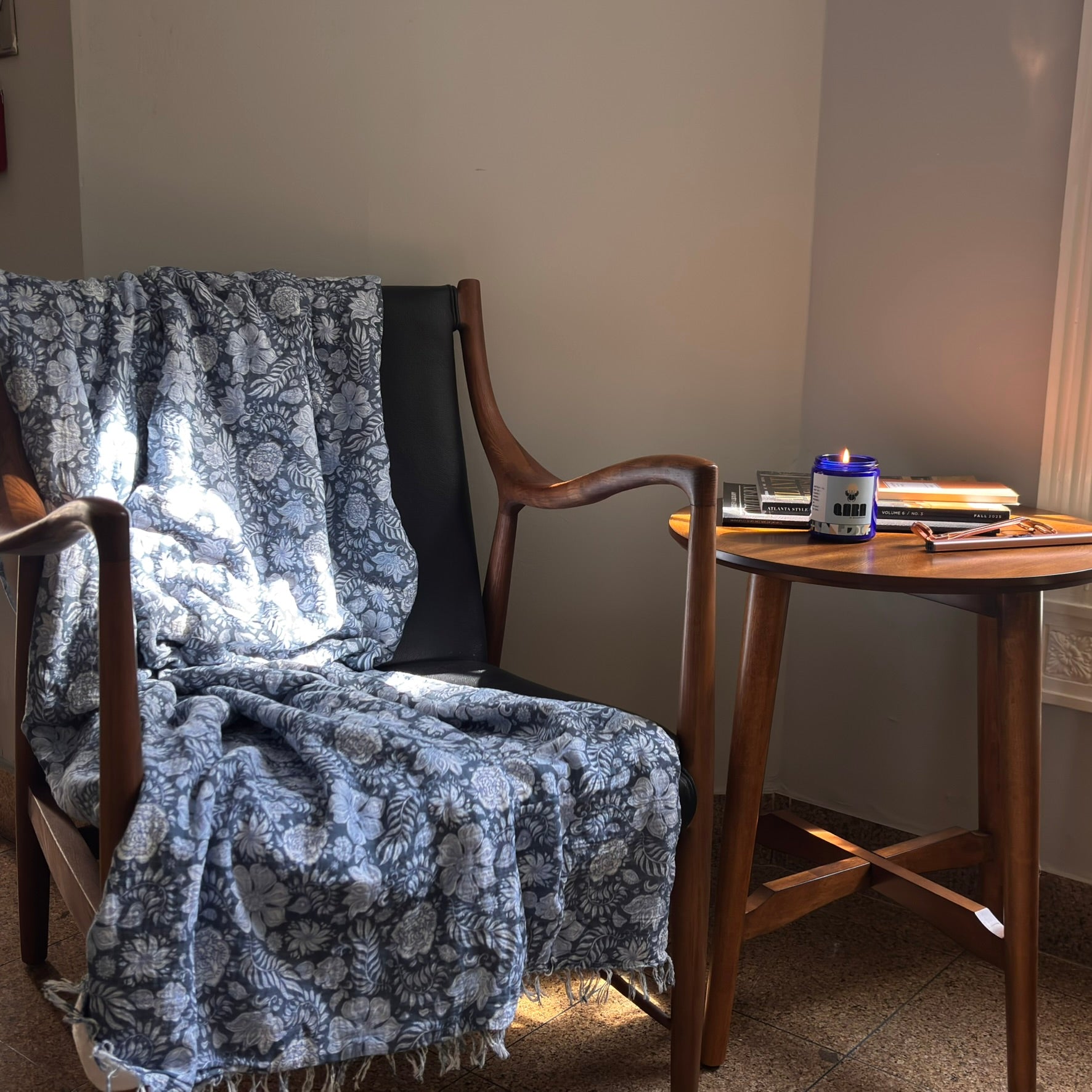 Chair with a blue floral blanket draped over it next to a wooden table with a can and notebook. QARA DEW scented candle fresh and flowery scent luxurious scent natural ingredients and lab tested fragrance oil front cobalt blue jar