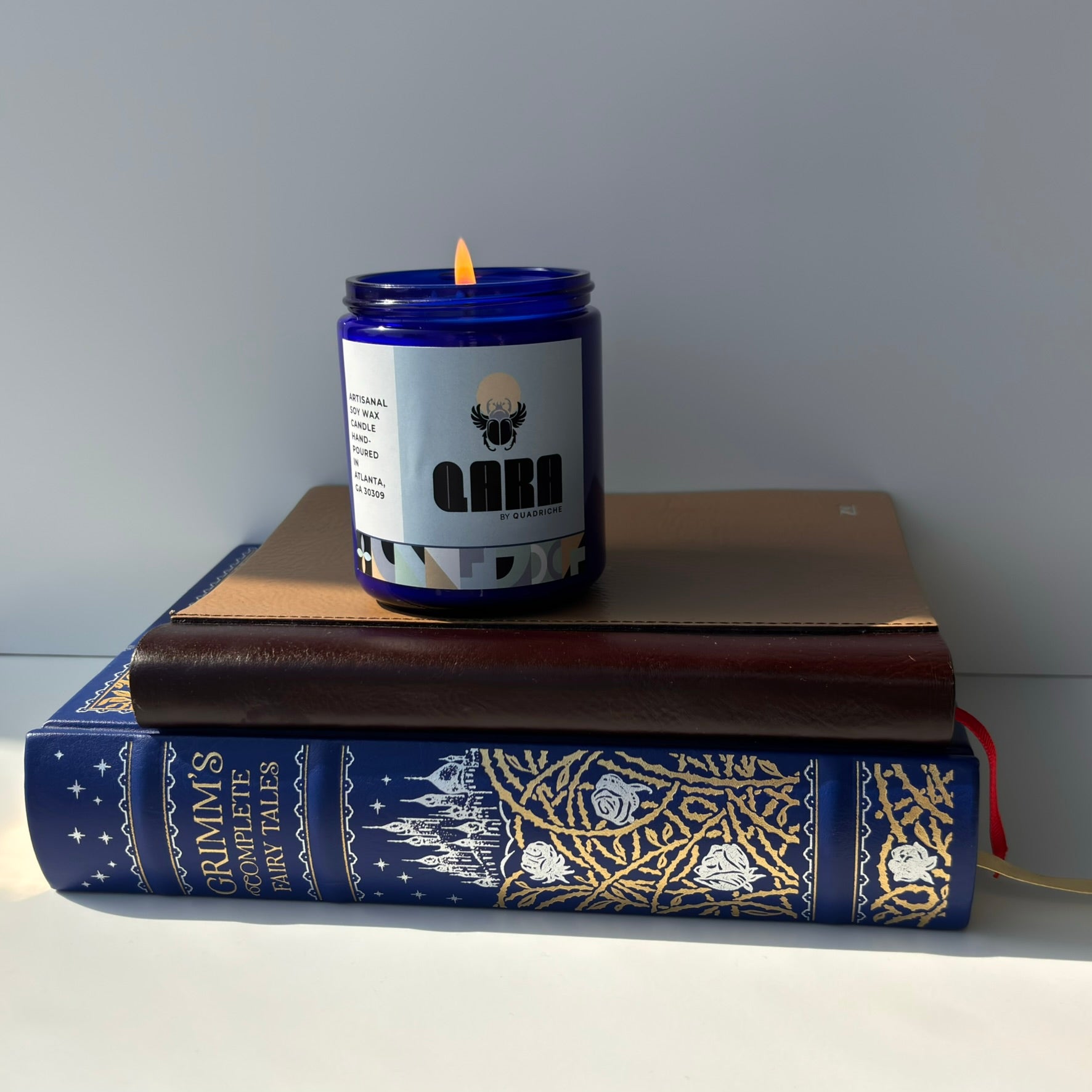 Candle with a label on top of two books against a plain background.  QARA DEW scented candle fresh and flowery scent luxurious scent natural ingredients and lab tested fragrance oil front cobalt blue jar