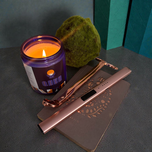 Candle in a purple jar with a lit wick, next to a rose gold lighter and decorative metal tray on a dark surface.