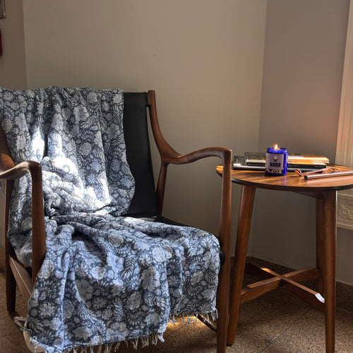 Chair with a floral blanket draped over it next to a small wooden table with a candle and books. QARA IVORY ORIS scented candle Violet Fig  flowery scent luxurious scent natural ingredients and lab tested fragrance oil front cobalt blue jar
