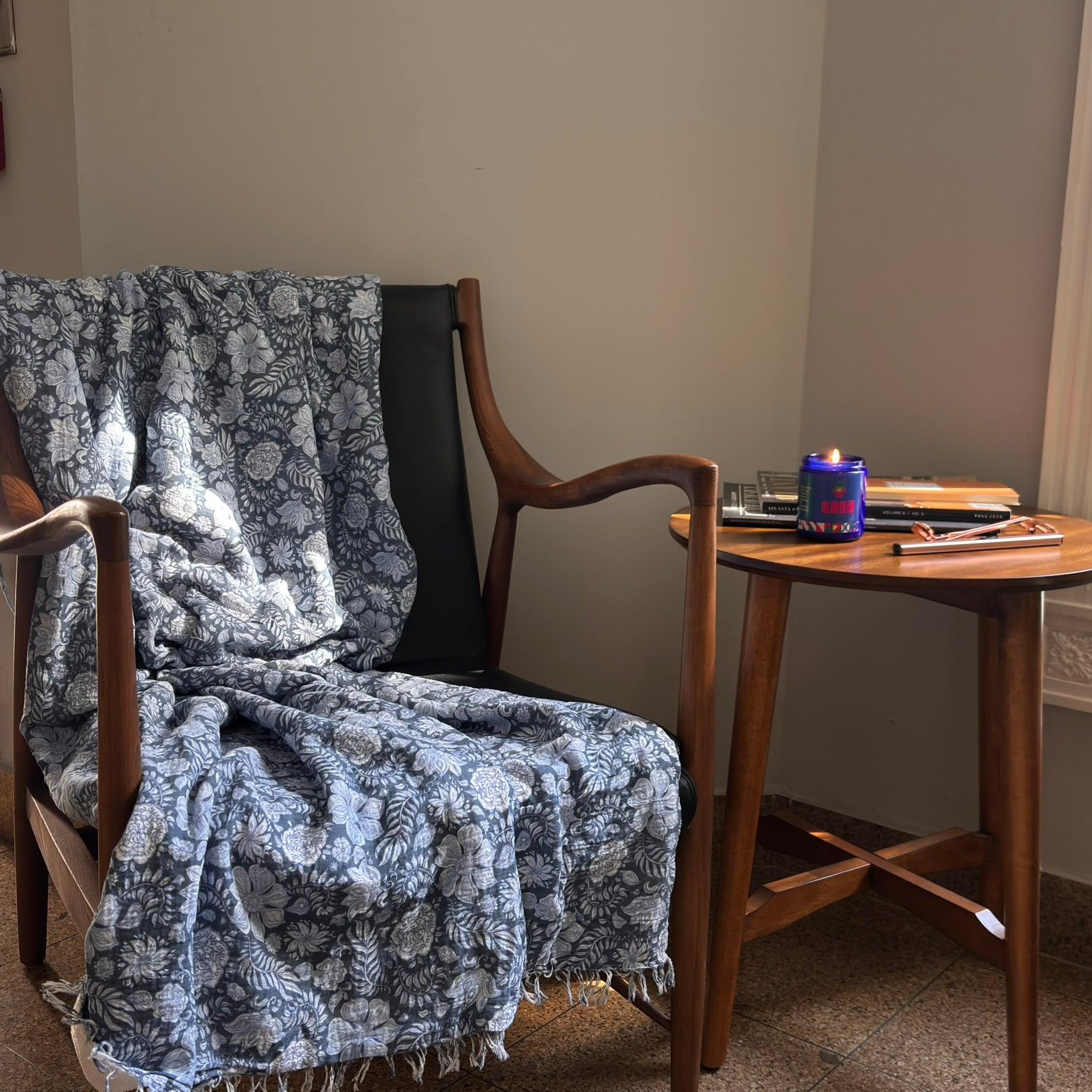 Wooden armchair with a floral-patterned blanket draped over it, next to a small wooden table with a candle and books. QARA Seventh Bloom Jasmine flower lab tested candle fragrance 