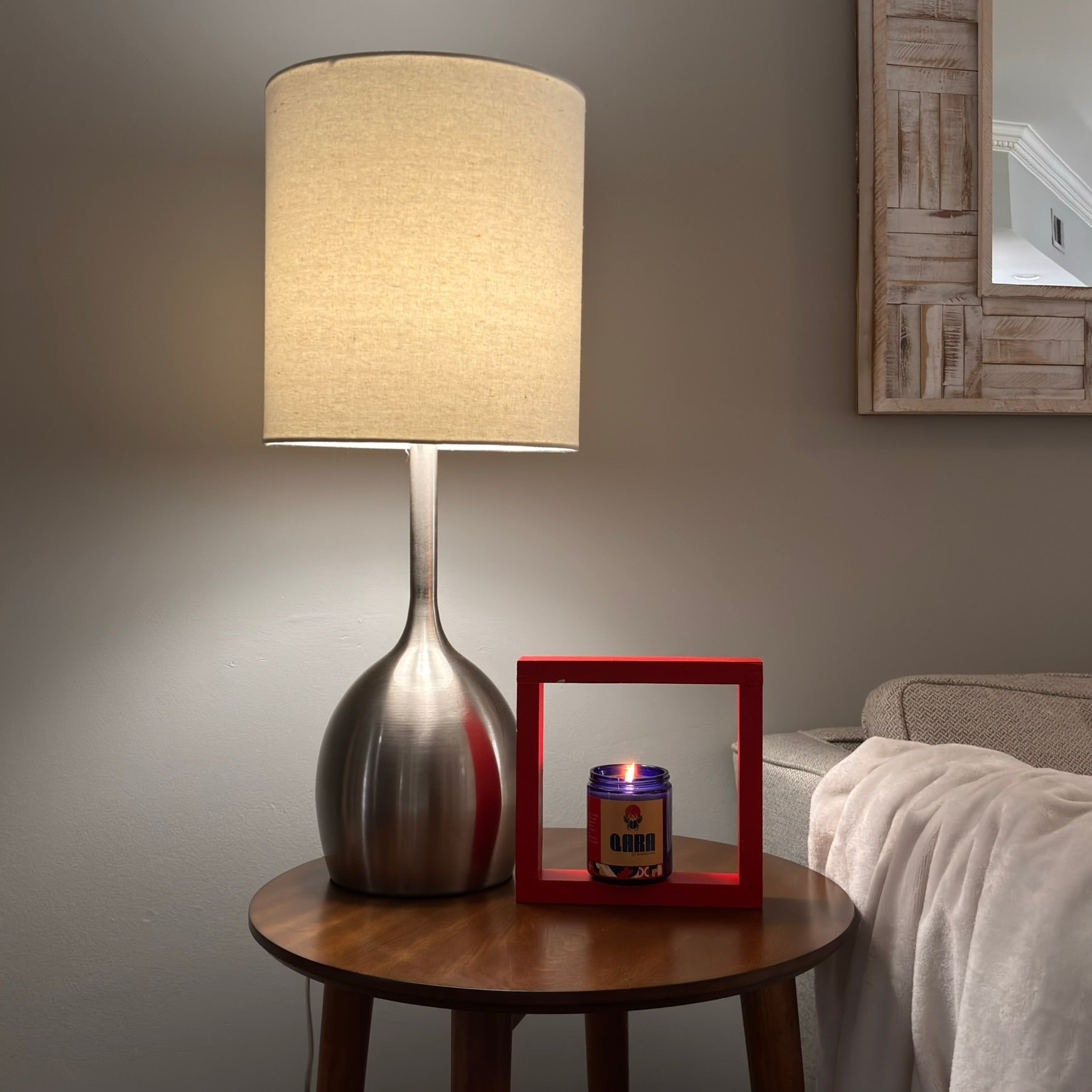 Table lamp on a wooden side table with a candle and red frame in a room setting.