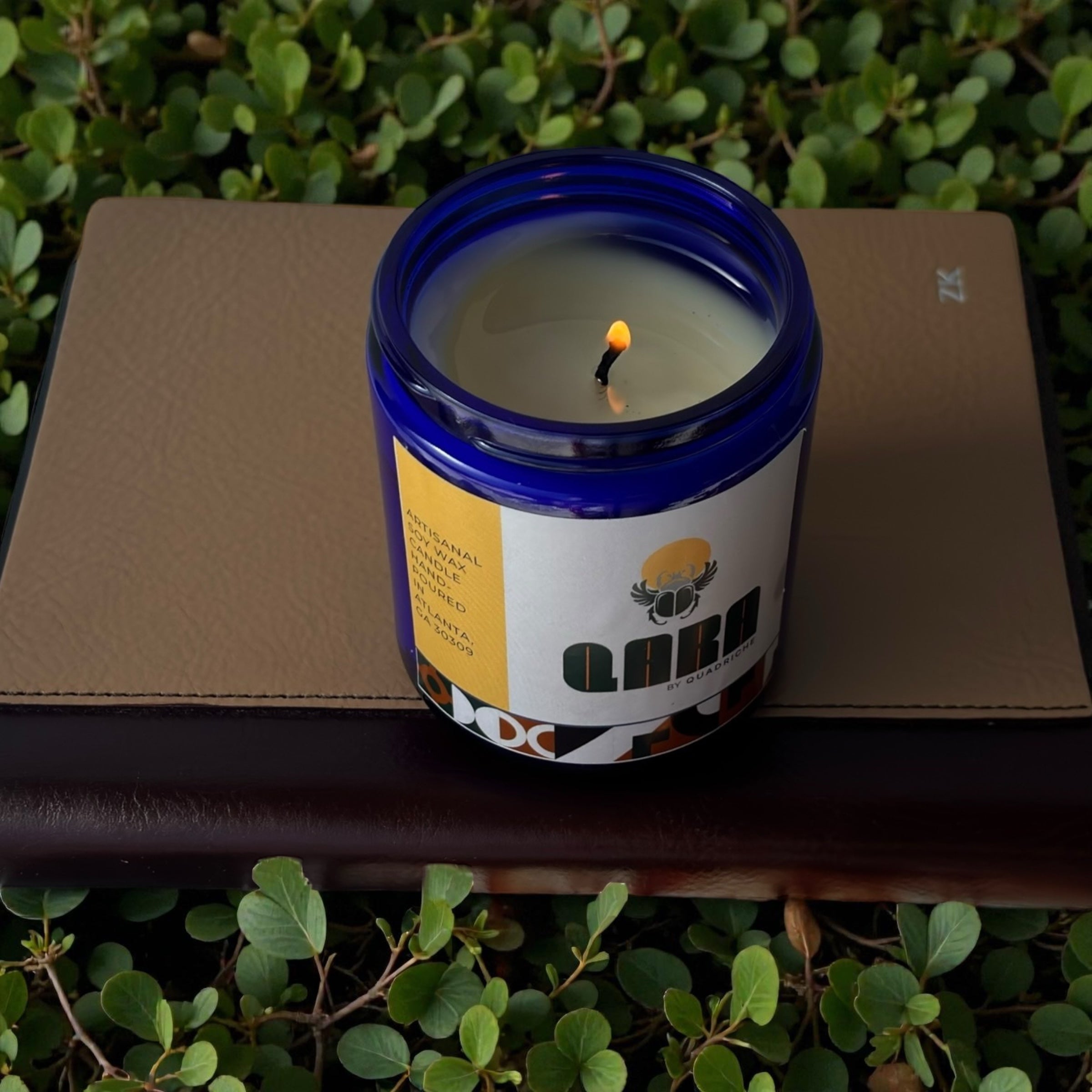Candle in a blue container with a visible flame, placed on a brown surface with green foliage in the background.