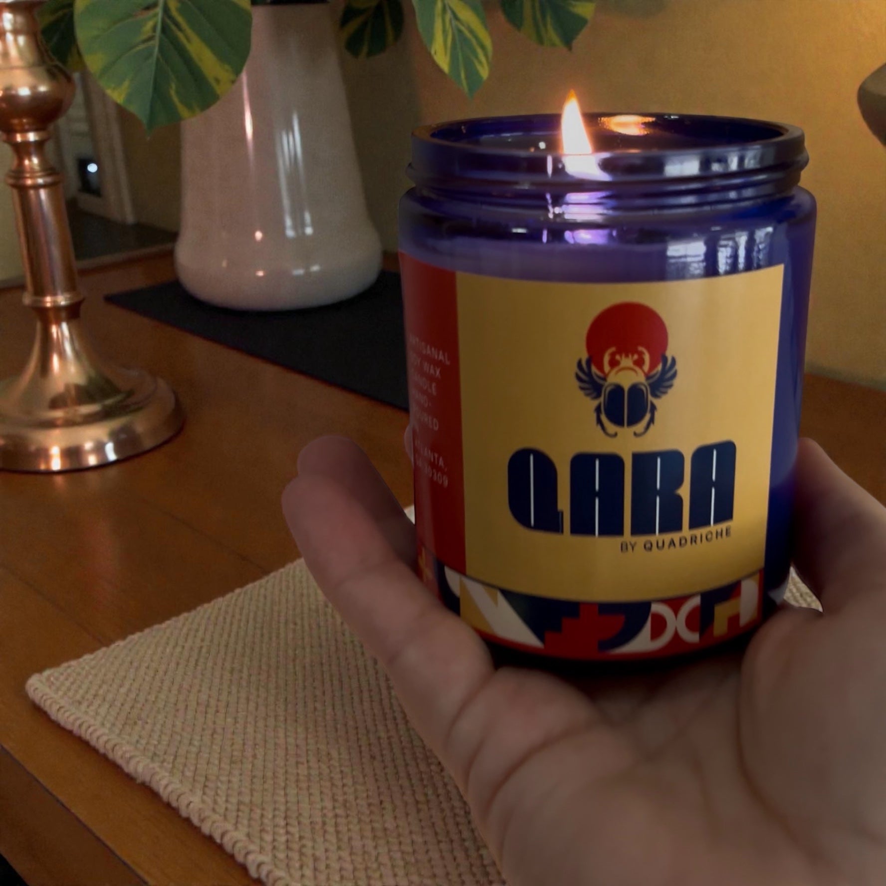 Candle in a colorful container labeled 'Qara' by Quadriche held by a hand on a wooden surface and linen placemat